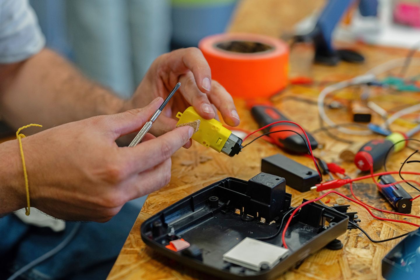 Hände, die mit einem Schraubenzieher an einem kleinen Getriebemotor arbeiten, daneben liegen Elektroschrott und Werkzeuge